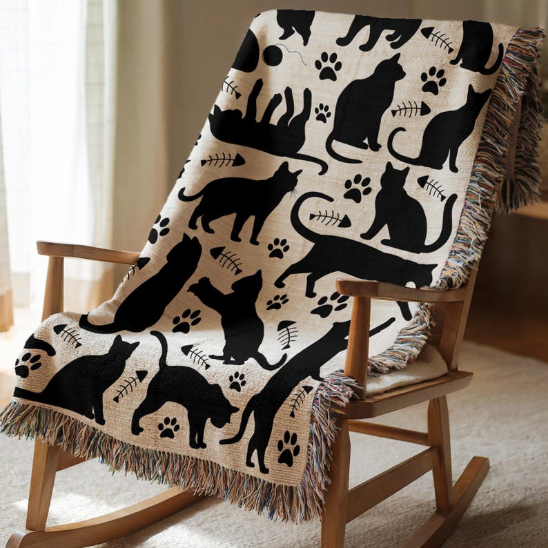May include: A beige throw blanket with a repeating pattern of black cat silhouettes, paw prints, and fish bones. The blanket is draped over a wooden rocking chair and has a fringed edge.