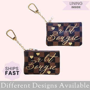 May include: Two small brown and black checkered coin purses with gold-colored hardware and the phrase "a bit Bougie" in gold script. Each purse has gold heart accents and a gold chain with a clasp. The text "Different Designs Available" is at the bottom.