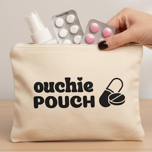 Op de afbeelding: Een beige canvas etui met de tekst "ouchie POUCH" in zwart, een hartje en een pil-afbeelding. De etui bevat een witte spuitfles en blisterverpakkingen met pillen. Een hand reikt in de etui.