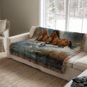 May include: A woven throw blanket featuring a painting of three brown horses running through water. The blanket has a fringed edge with shades of brown, blue, and tan. The blanket is draped over a white sofa.