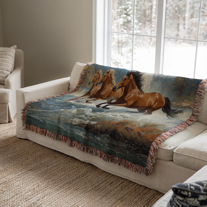 May include: A woven throw blanket featuring a painting of three brown horses running through water. The blanket has a fringed edge with shades of brown, blue, and tan. The blanket is draped over a white sofa.
