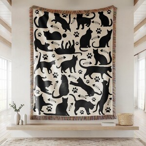 May include: A cream-colored throw blanket featuring a repeating pattern of black cat silhouettes in various poses, paw prints, and fish bones. The blanket has a fringed edge and is displayed on a wooden shelf.