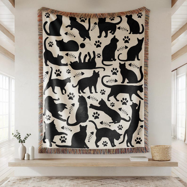 May include: A cream-colored throw blanket featuring a repeating pattern of black cat silhouettes in various poses, paw prints, and fish bones. The blanket has a fringed edge and is displayed on a wooden shelf.