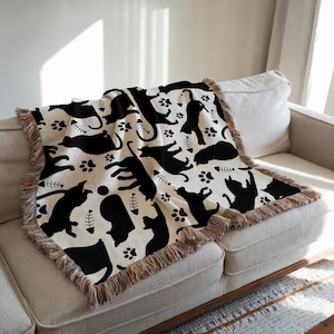 May include: A cream-colored throw blanket featuring a repeating pattern of black cat silhouettes, paw prints, and fish bones. The blanket has a fringed edge and is draped over a beige sofa. The design creates a playful and whimsical aesthetic.