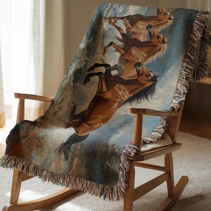 May include: A woven throw blanket draped over a wooden rocking chair. The blanket features a detailed illustration of several brown horses in motion against a blue and white background. The blanket has a fringed edge.