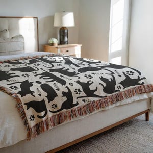 May include: A beige throw blanket with a repeating pattern of black cat silhouettes, paw prints, and fish bones. The blanket has a fringed edge with a mix of colors and is draped over a bed.