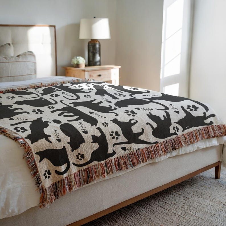 May include: A beige throw blanket with a repeating pattern of black cat silhouettes, paw prints, and fish bones. The blanket has a fringed edge with a mix of colors and is draped over a bed.