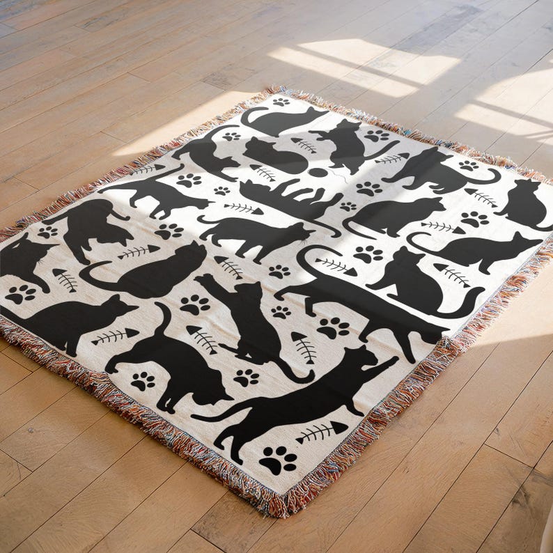 May include: A cream-colored throw blanket featuring a repeating pattern of black cat silhouettes, paw prints, and fish bones. The blanket has a fringed edge and is made of a soft, woven material. The design is playful and suitable for cat lovers.