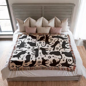 May include: A cream-colored throw blanket with a black cat pattern, paw prints, and fish bones. The blanket has a fringed edge and is displayed on a bed with pillows. The bed frame is gray.