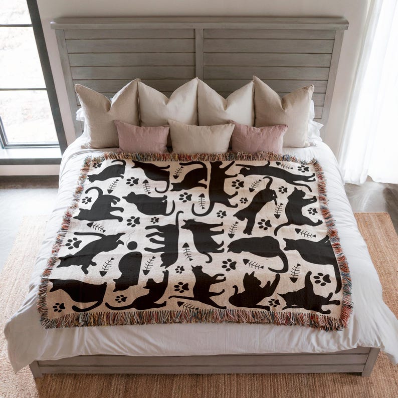 May include: A cream-colored throw blanket with a black cat pattern, paw prints, and fish bones. The blanket has a fringed edge and is displayed on a bed with pillows. The bed frame is gray.
