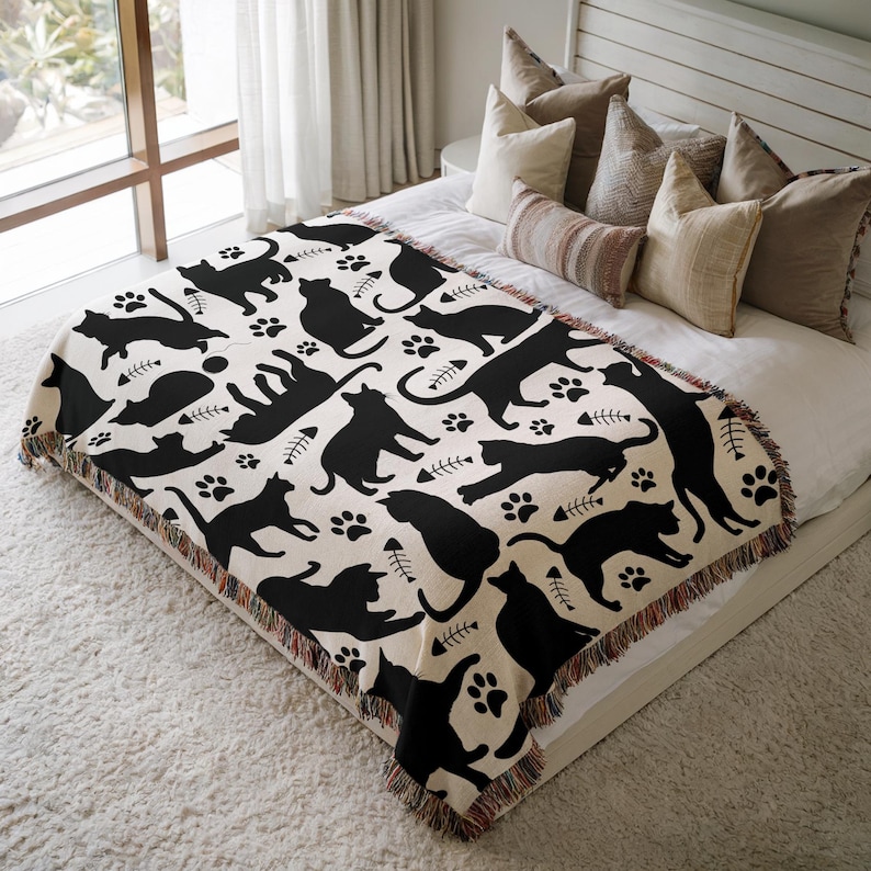 May include: A cream-colored throw blanket featuring a repeating pattern of black cat silhouettes, paw prints, and fish bones. The blanket has a fringed edge and is displayed on a bed with pillows.