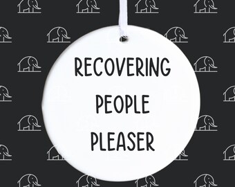 Może przedstawiać: Biała ozdoba ceramiczna z napisem "RECOVERING PEOPLE PLEASER" czarnymi literami.