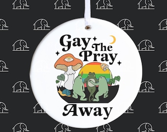 May include: A round, white ceramic ornament with a colourful graphic. The design features a mushroom with an eye, frogs, and the text "Gay the Pray Away." A white ribbon is attached for hanging. The background is black with white elephant shapes.