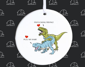 May include: A white ceramic ornament with a humorous illustration of two Tyrannosaurus Rex dinosaurs. One is green, the other is blue. Text includes "I'M FUCKING TRYING!" and "PULL MY HAIR!" with heart symbols. A white ribbon is attached.