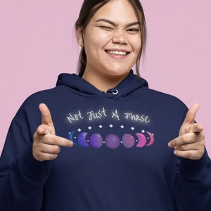 7 phases of the moon in a gradient of the bisexual pride colors: blue, purple, and magenta, with the words Not Just a Phase in a handwritten font above. It is demoed the front of a navy pullover hoodie on an asian woman winking and doing finger guns.