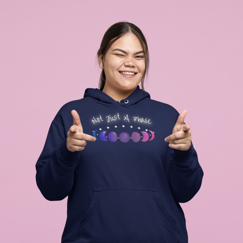 7 phases of the moon in a gradient of the bisexual pride colors: blue, purple, and magenta, with the words Not Just a Phase in a handwritten font above. It is demoed the front of a navy pullover hoodie on an asian woman winking and doing finger guns.