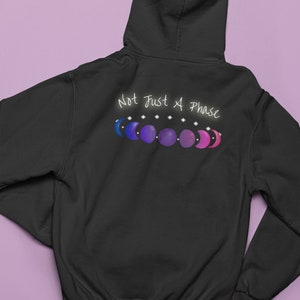 7 phases of the moon in a gradient of the bisexual pride colors: blue, purple, and magenta, with the words Not Just a Phase in a handwritten font above. It is demoed on the back of a black zip-up hoodie laid flat on a purple background.