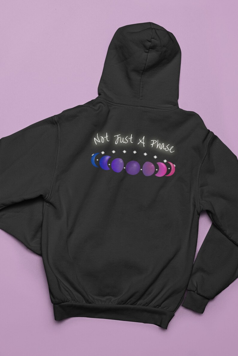 7 phases of the moon in a gradient of the bisexual pride colors: blue, purple, and magenta, with the words Not Just a Phase in a handwritten font above. It is demoed on the back of a black zip-up hoodie laid flat on a purple background.