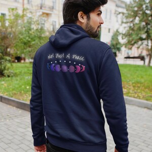 7 phases of the moon in a gradient of the bisexual pride colors: blue, purple, and magenta, with the words Not Just a Phase in a handwritten font above. It is demoed on the back of a navy zip-up hoodie on a feminine man with dark thick hair.