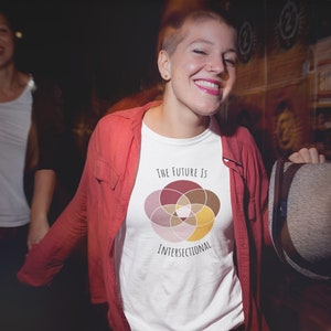 Five interlocking circles of color with white borders arranged in a flower , in shades of pink, maroon, brown, and canary yellow. Black text above and below says The Future Is Intersectional. It is on a white T-shirt worn by a sassy queer woman.