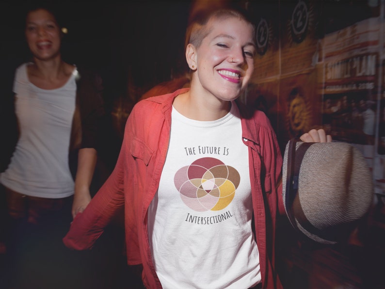 Five interlocking circles of color with white borders arranged in a flower , in shades of pink, maroon, brown, and canary yellow. Black text above and below says The Future Is Intersectional. It is on a white T-shirt worn by a sassy queer woman.