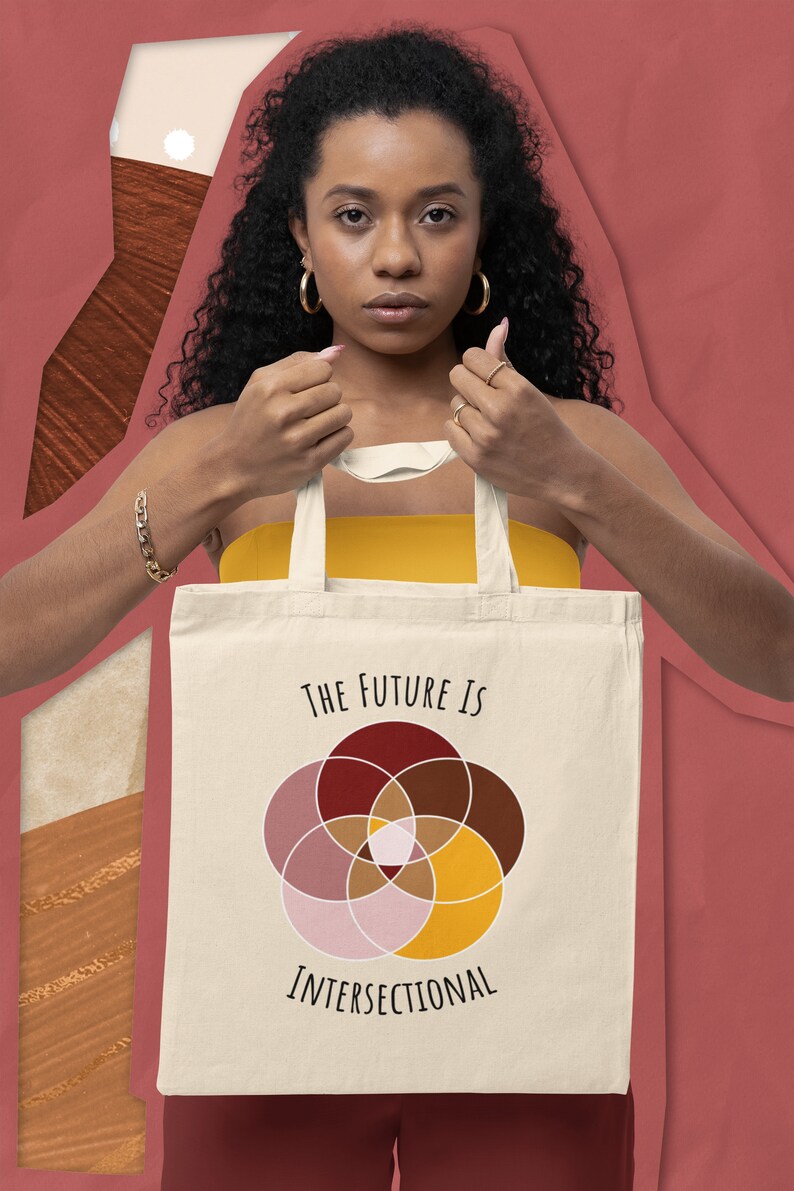 A woman of color holding a beige canvas bag with five interlocking white-bordered circles, arranged in a flower in shades of pink, maroon, brown, and canary yellow, and black text above and below that says The Future Is Intersectional, on it.