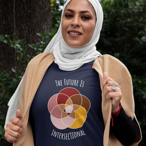 Five interlocking circles of color with white borders arranged in a flower , in shades of pink, maroon, brown, and canary yellow. White text above and below says The Future Is Intersectional. It is on a navy T-shirt worn by a muslim woman.