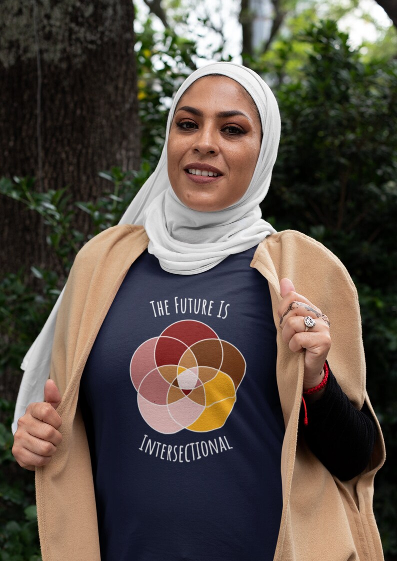 Five interlocking circles of color with white borders arranged in a flower , in shades of pink, maroon, brown, and canary yellow. White text above and below says The Future Is Intersectional. It is on a navy T-shirt worn by a muslim woman.