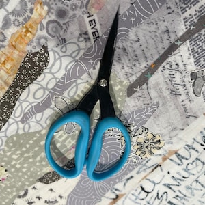 May include: Black sewing scissors with bright blue handles rest on a patchwork of fabric. The fabric features a mix of gray, white, and tan patterns, including boots, floral designs, and text. The scissors are open, ready for use in a sewing or crafting project.