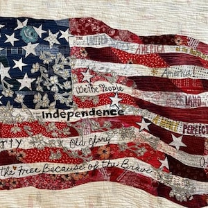May include: A quilted American flag with a waving design. The flag features red and white stripes, a blue field with white stars, and text including "Liberty," "Independence," and "Land of the Free." The flag is made of fabric with floral patterns.