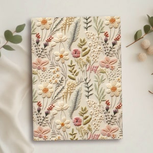May include: A rectangular notebook with an embroidered floral design. The cream-colored cover features a repeating pattern of flowers and foliage in shades of pink, yellow, green, and beige. The design is detailed and textured.