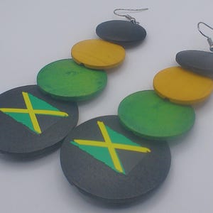 Può includere: Un paio di orecchini pendenti con un design ispirato alla bandiera giamaicana. Gli orecchini hanno una serie di dischi di legno in nero, giallo e verde, con il disegno a croce della bandiera giamaicana sui dischi più grandi. Gli orecchini hanno ganci color argento.