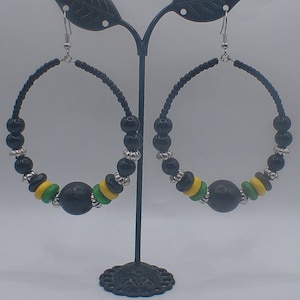 Può includere: Un paio di grandi orecchini a cerchio con perline e dettagli neri. Gli orecchini presentano una combinazione di perline nere, gialle e verdi, con dettagli in metallo color argento. Gli orecchini sono esposti su un supporto in metallo nero.