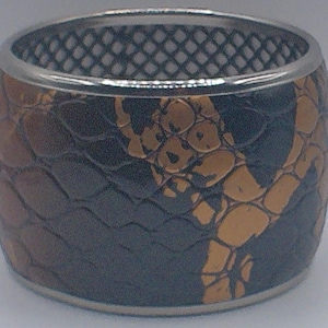 May include: A wide, cylindrical ring with a textured, reptile-like pattern in shades of brown, black, and gold. The ring has a silver-colored band at the top and bottom. The interior has a black, grid-like pattern.