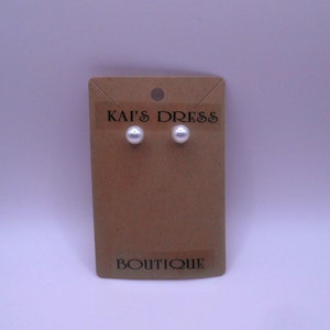 May include: A pair of white pearl stud earrings on a brown card with the text "Kai's Dress Boutique".
