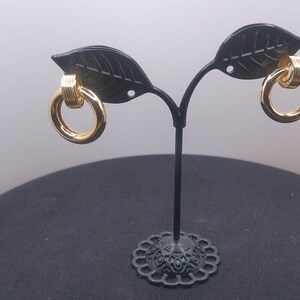 Può includere: Un paio di orecchini a cerchio dorati con un design intrecciato. Gli orecchini sono esposti su un supporto nero con bracci a forma di foglie.