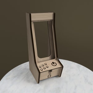 Op de afbeelding: Een houten telefoonstandaard in arcade-stijl met een uitsparing voor een telefoon en een bedieningspaneel met knoppen en een aan/uit-knop. De standaard is gemaakt van lichtbruin hout met donkerbruine afwerking.