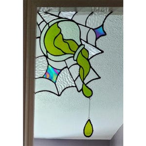 Può includere: Pannello di finestra in vetro colorato con un design verde e bianco. L'opera d'arte raffigura un vaso con liquido verde che scorre, con vetro iridescente e contorni neri. Una goccia verde è appesa sotto.