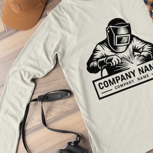 Op de afbeelding: Een longsleeve t-shirt met een zwart-witte illustratie van een lasser die een helm draagt en een stuk metaal last. De tekst "COMPANY NAME" is onder de illustratie gedrukt.