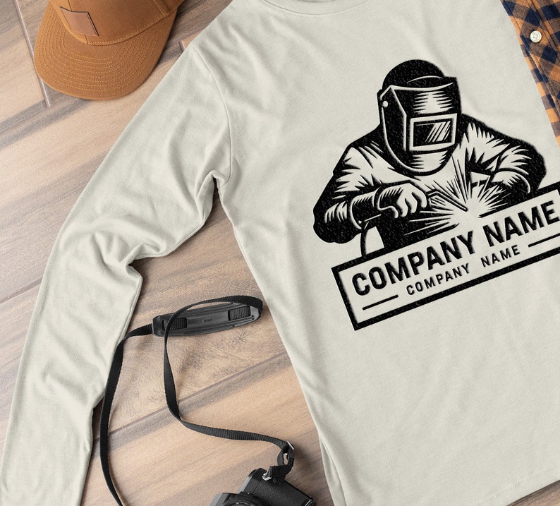 Op de afbeelding: Een longsleeve t-shirt met een zwart-witte illustratie van een lasser die een helm draagt en een stuk metaal last. De tekst "COMPANY NAME" is onder de illustratie gedrukt.