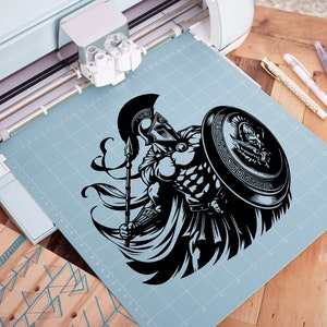 K&ouml;nnte beinhalten: Eine Schwarzwei&szlig;illustration eines griechischen Kriegers, der einen Speer und einen Schild h&auml;lt. Der Krieger tr&auml;gt einen Helm und eine R&uuml;stung. Die Illustration befindet sich auf einer blauen Schneidematte.