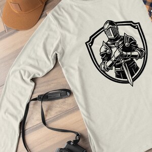 May include: A long-sleeved, light grey t-shirt with a black and white graphic of a knight in armour holding a sword, standing inside a shield.
