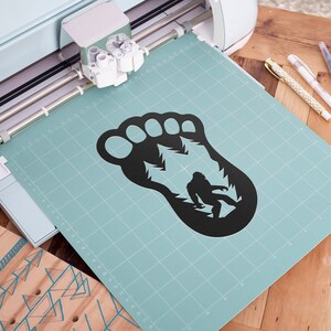 K&ouml;nnte beinhalten: Eine schwarze Silhouette eines Bigfoot-Fu&szlig;abdrucks mit einer kleineren Silhouette von Bigfoot innerhalb des Fu&szlig;abdrucks. Der Fu&szlig;abdruck befindet sich auf einer hellblauen Schneidematte mit einem Gittermuster.