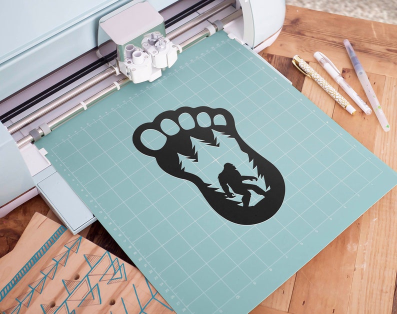 K&ouml;nnte beinhalten: Eine schwarze Silhouette eines Bigfoot-Fu&szlig;abdrucks mit einer kleineren Silhouette von Bigfoot innerhalb des Fu&szlig;abdrucks. Der Fu&szlig;abdruck befindet sich auf einer hellblauen Schneidematte mit einem Gittermuster.