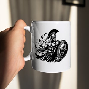 K&ouml;nnte beinhalten: Wei&szlig;e Keramiktasse mit einer Schwarzwei&szlig;illustration eines spartanischen Kriegers, der einen Speer und einen Schild h&auml;lt. Der Krieger tr&auml;gt einen Helm und eine R&uuml;stung.
