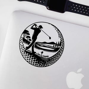 May include: Black and white golf-themed sticker showing a golfer in action within a golf ball design. The scene includes a golf course with trees, hills, and a tee. The sticker is placed on a white surface.