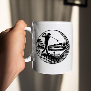 May include: White ceramic mug with a black and white golf design. The design shows a golfer swinging a club within a golf ball-shaped frame, with a golf course landscape. The mug has a curved handle.