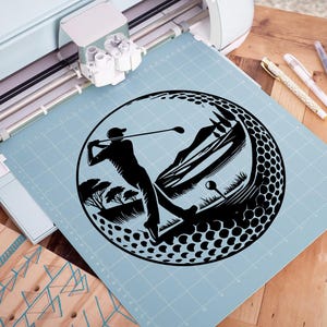 May include: Black silhouette of a golfer swinging a club, set within a golf ball design. The image includes a mountain and tree landscape. The design is on a light blue cutting mat, with a Cricut machine in the background.