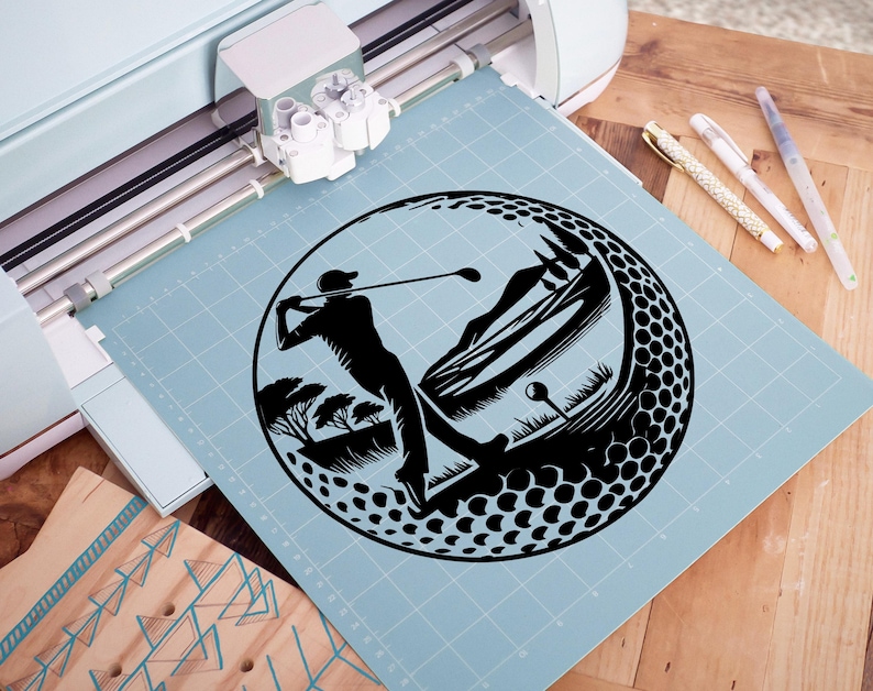May include: Black silhouette of a golfer swinging a club, set within a golf ball design. The image includes a mountain and tree landscape. The design is on a light blue cutting mat, with a Cricut machine in the background.
