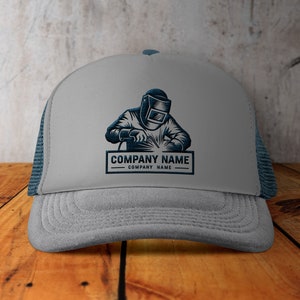 Op de afbeelding: Grijze truckercap met een blauw mesh achterpaneel. De voorkant van de cap heeft een zwart-witte illustratie van een lasser met een lasmasker en de tekst "COMPANY NAME COMPANY NAME".